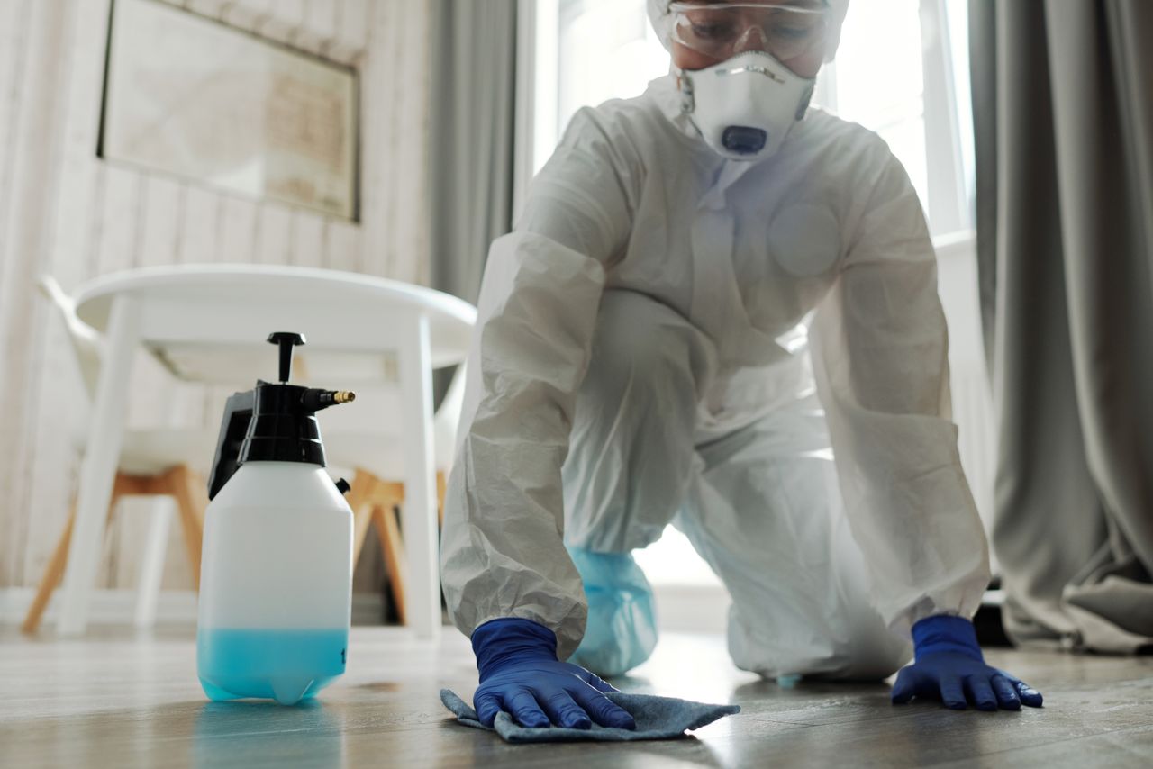 services-03 A person wearing protective equipment cleans an indoor floor using disinfectant spray and a cloth.
